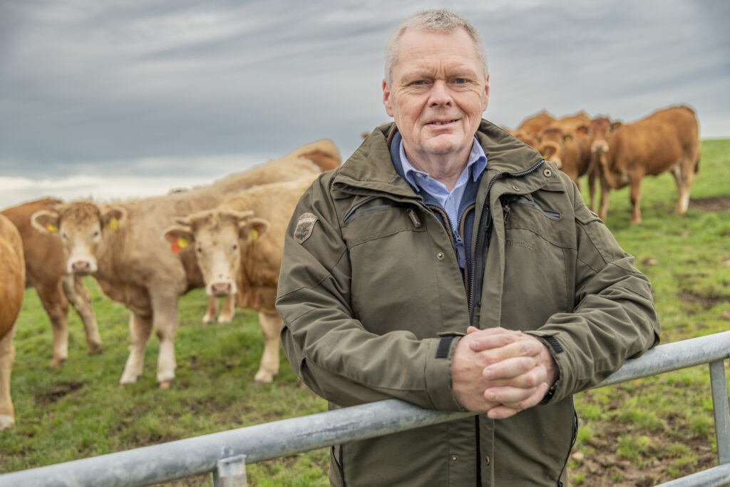 The president of the NFU Scotland, Andrew Connon pictured leaning up against a metal gate leading into a field with cattle.
